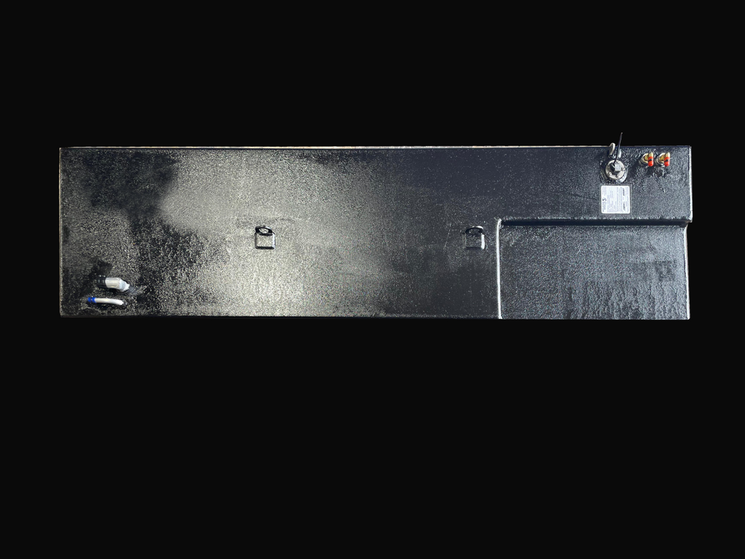 A direct top-down photo of a long, dark, textured boat fuel tank with a stepped design at one end, showing its various top-mounted fittings, D-rings, and specification plate against a black background.