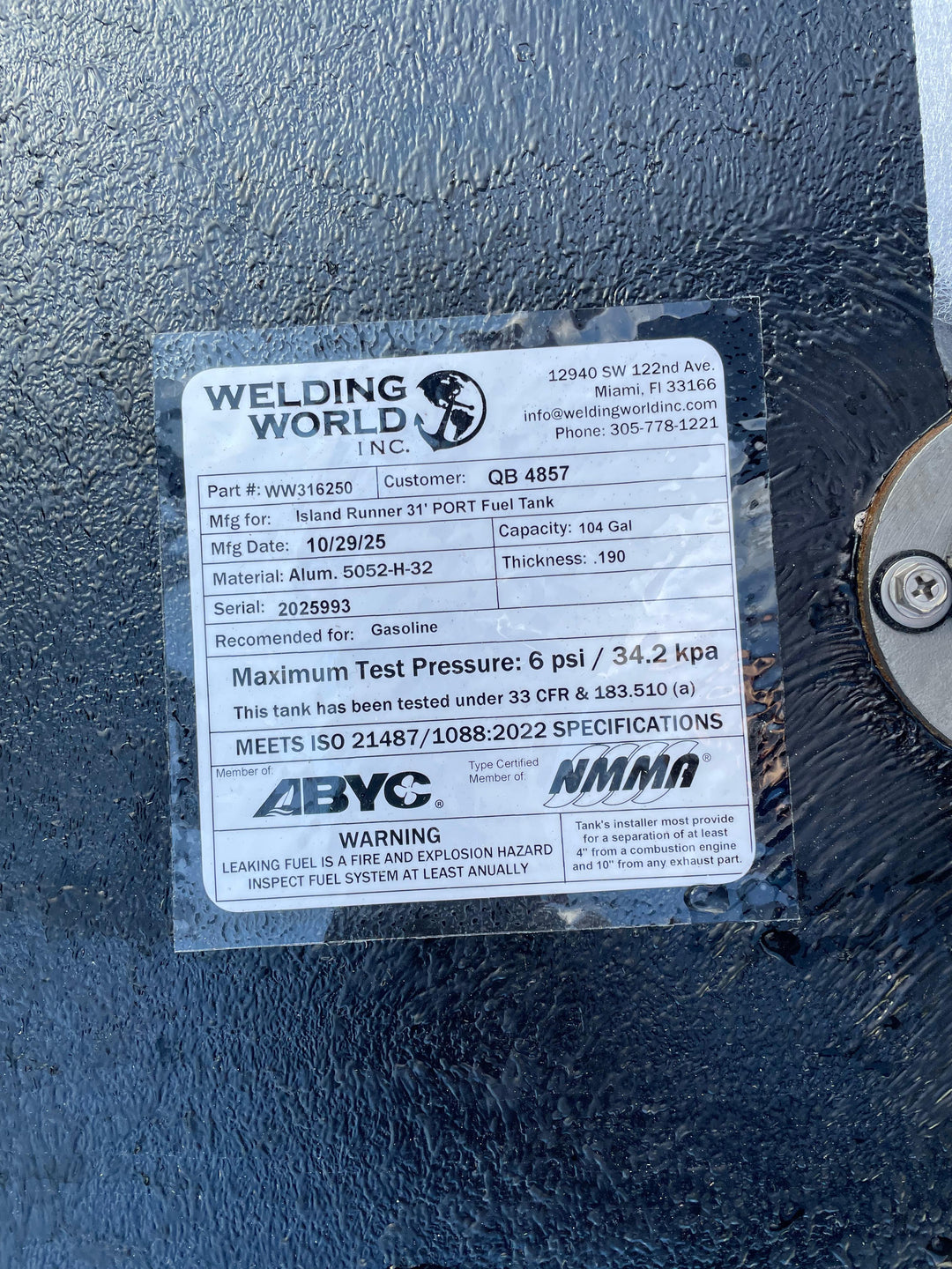 A close-up of a white specification label, shown upside down, on a dark textured boat fuel tank. The Welding World Inc. label indicates it is for an "Island Runner 31' PORT Fuel Tank" with a 104-gallon capacity, part number WW316250.