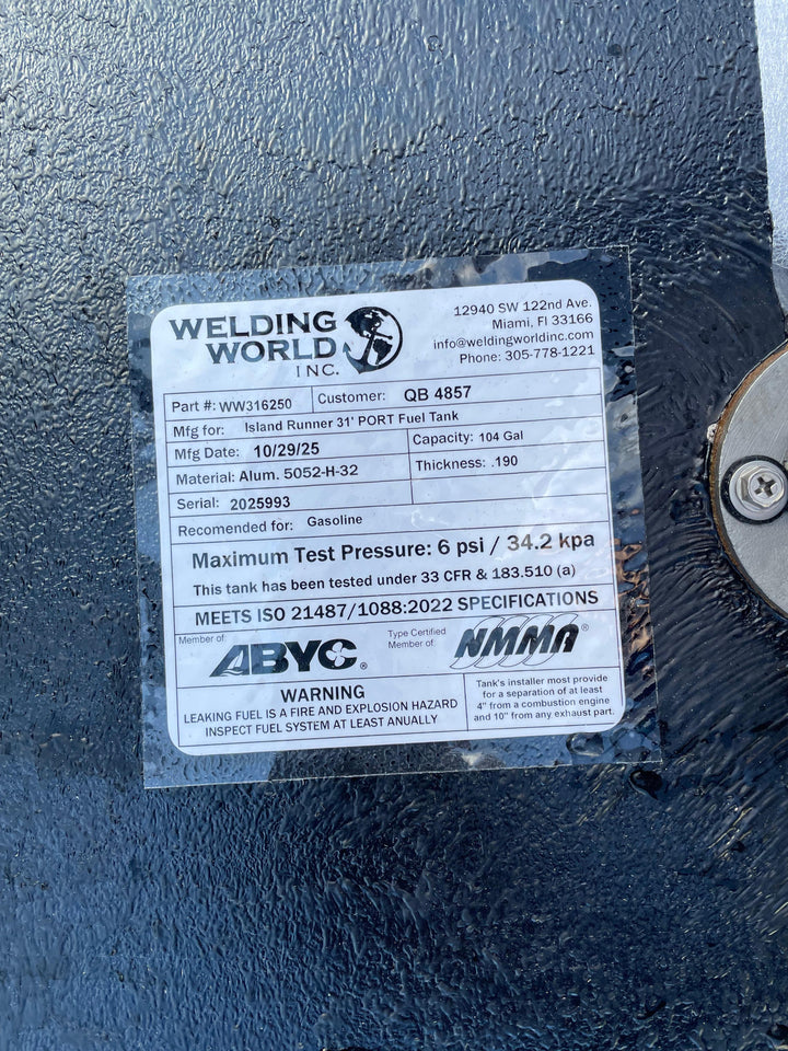 A close-up of a white specification label, shown upside down, on a dark textured boat fuel tank. The Welding World Inc. label indicates it is for an "Island Runner 31' PORT Fuel Tank" with a 104-gallon capacity, part number WW316250.