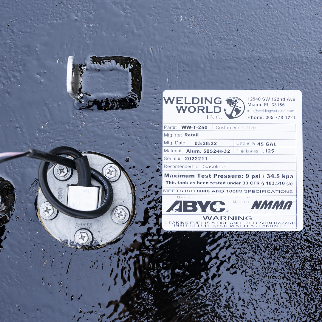 A white product label from "Welding World Inc." affixed to a wet, textured black surface. The label provides details for a "WW-T-250" fuel tank, indicating it's for retail, manufactured on 03/28/22, made of Alum. 5052-H-32 with a thickness of .125, and has a capacity of 45 gallons. It also lists the serial number 2022211, states it's recommended for gasoline, and has a maximum test pressure of 9 psi / 34.5 kpa, meeting ISO 8846 and 10088 specifications. A circular metal component with wires and screws