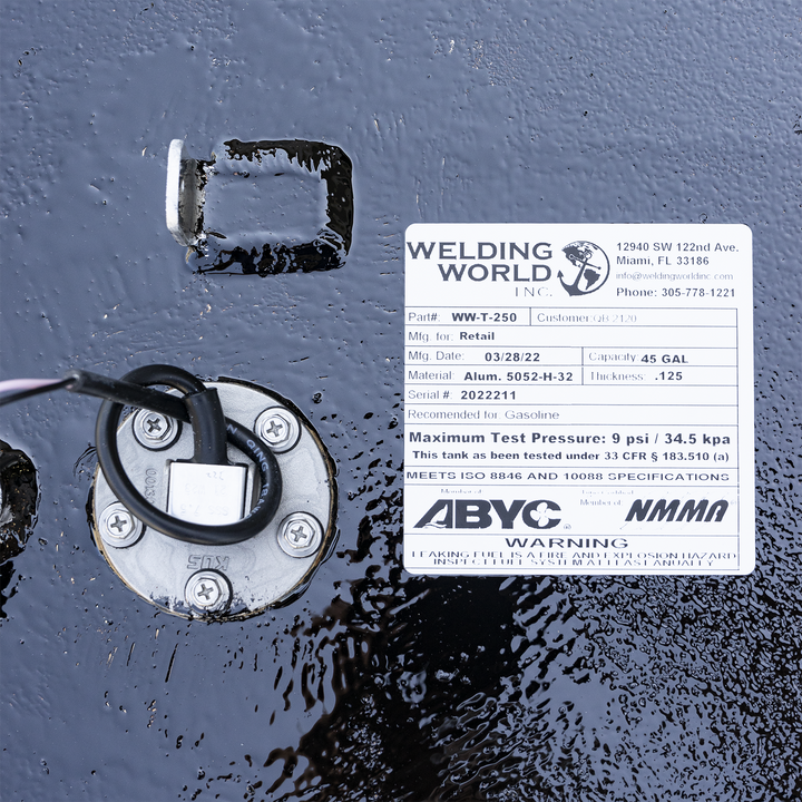 A white product label from "Welding World Inc." affixed to a wet, textured black surface. The label provides details for a "WW-T-250" fuel tank, indicating it's for retail, manufactured on 03/28/22, made of Alum. 5052-H-32 with a thickness of .125, and has a capacity of 45 gallons. It also lists the serial number 2022211, states it's recommended for gasoline, and has a maximum test pressure of 9 psi / 34.5 kpa, meeting ISO 8846 and 10088 specifications. A circular metal component with wires and screws