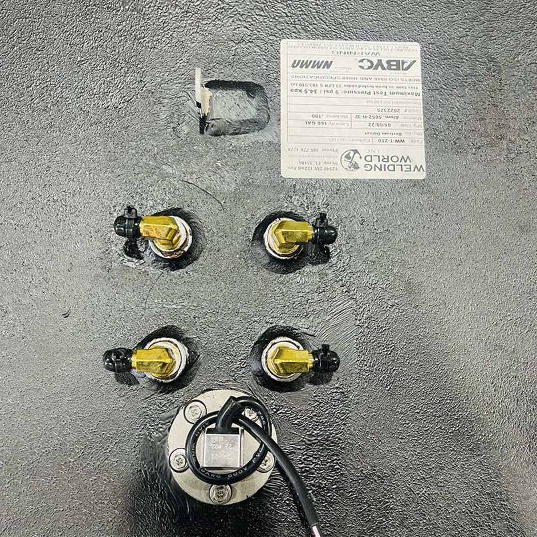 An overhead close-up view of a textured dark grey surface, likely a fuel tank, shows several brass fittings with black caps arranged in a square pattern. Below these fittings is a circular metallic plate secured by screws, with a black wire extending from its center. In the upper right corner, a white label with "WELDING WORLD INC." and other specifications is partially visible