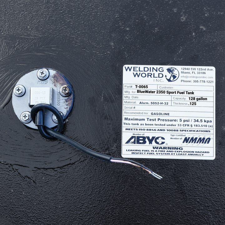 Close-up photograph of the Welding World Inc. certification label for a BlueWater 2350 Sport fuel tank, showing the 128-gallon capacity, .125" aluminum thickness, and ABYC/NMMA compliance stamps next to a KUS fuel sending unit.
