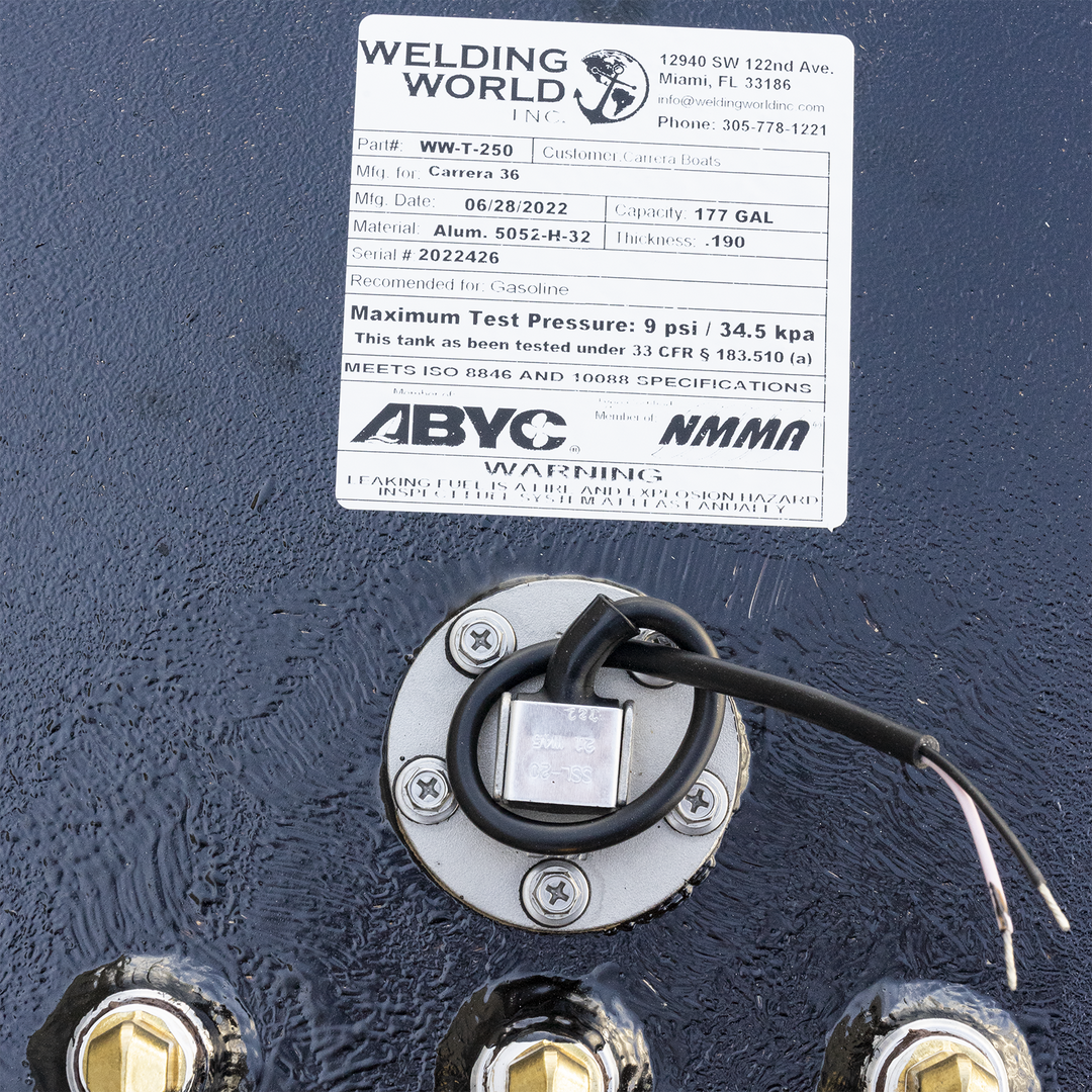 A close-up of a black, textured fuel tank with a white product label. The label, from Welding World Inc., details the tank as Part# WW-T-250, manufactured on 06/28/2022 for Carrera 36, with a capacity of 177 gallons, made from Alum. 5052-H-32 with a thickness of .190, and serial # 2022426. It's recommended for gasoline and has a maximum test pressure of 9 psi / 34.5 kpa, meeting ISO 8846 and 10088 specifications. Below the label, a circular silver-colored fitting with four screws, a black cable, and exposed