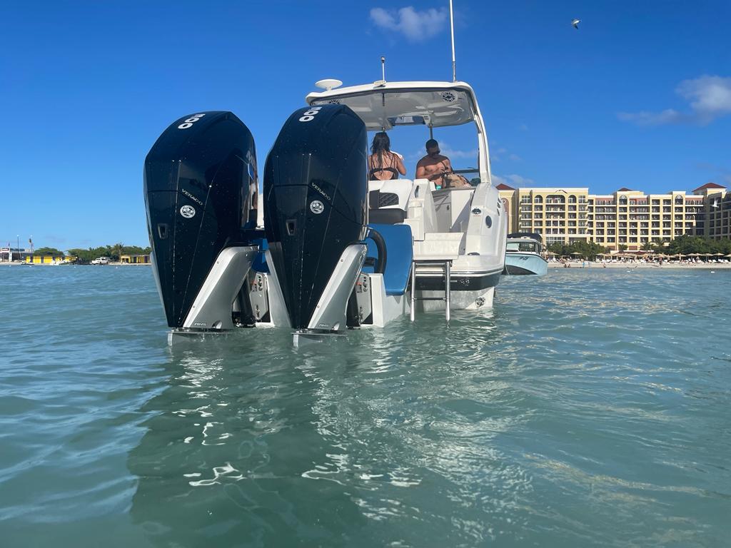 A white boat with two people on board, featuring a Chaparral 327 SSX with a custom high flotation twin outboard engine bracket, floating on calm blue water under a partly cloudy sky. In the background, there are buildings along the shoreline