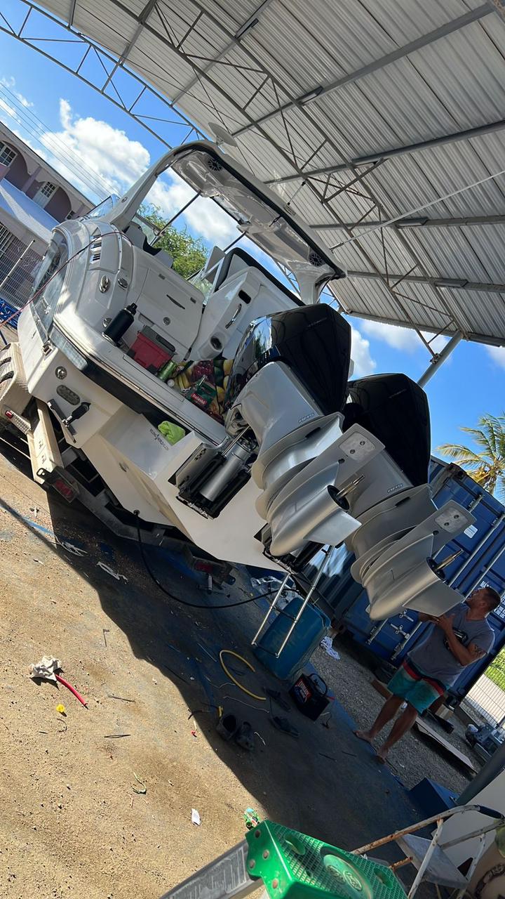 The back of a white boat with multiple outboard engines mounted on the stern, under a metal roofed structure, with a person standing nearby holding a device. The boat is out of the water, with tools and equipment on the ground around it