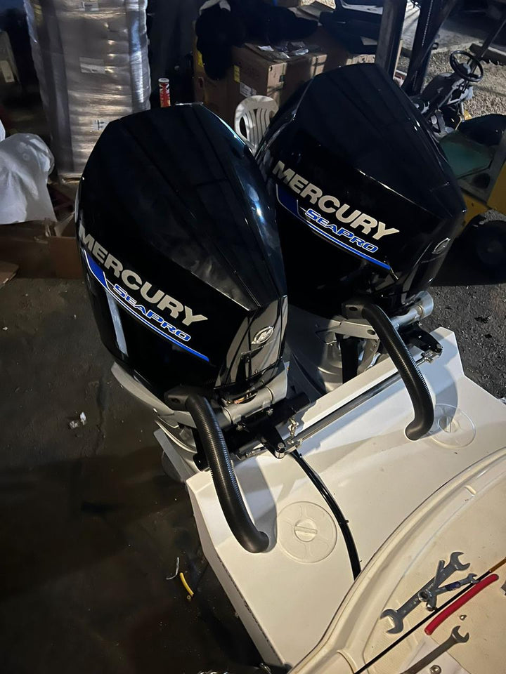 Two black Mercury SeaPro outboard engines mounted on a white boat, viewed from above in a workshop or storage area. The engines are positioned side by side, with hoses and mounting brackets visible. In the background, there are various tools, boxes, and equipment
