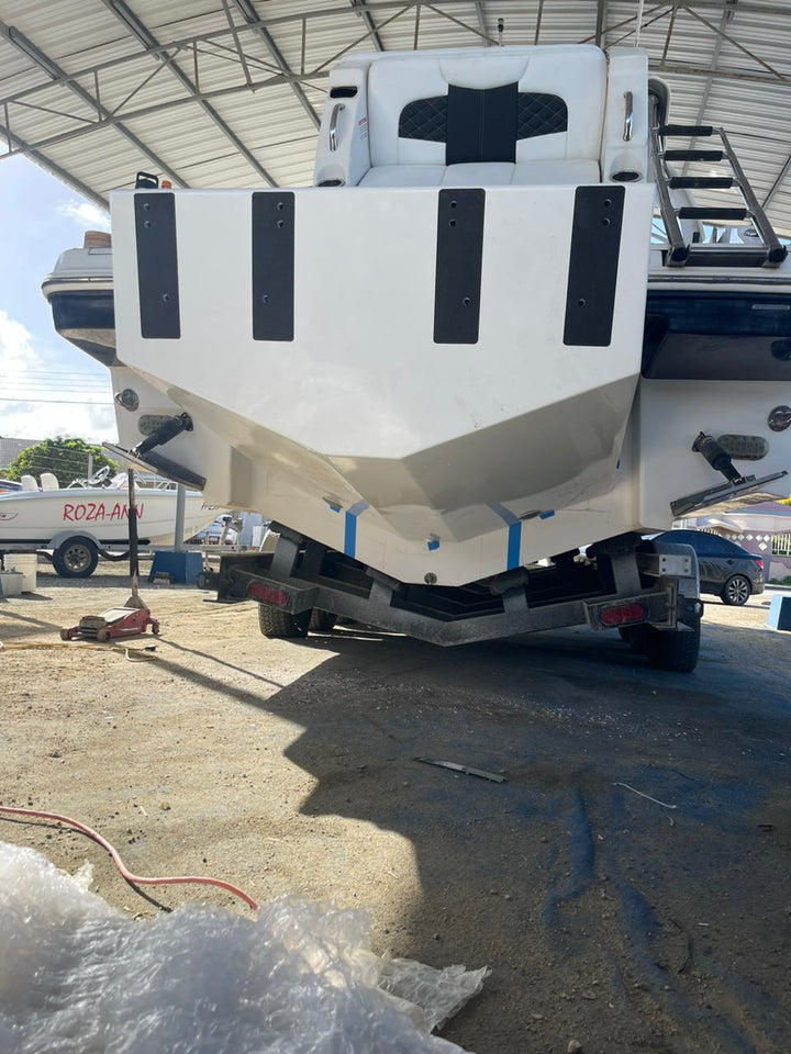 The front view of a white Chaparral 327 SSX boat with a custom high flotation twin outboard engine bracket, parked on a trailer under a metal canopy. The boat's bow features black accents and a seating area, and there are other boats and vehicles visible in the background. The boat is supported by a black trailer with red taillights, and the ground is a dirt surface