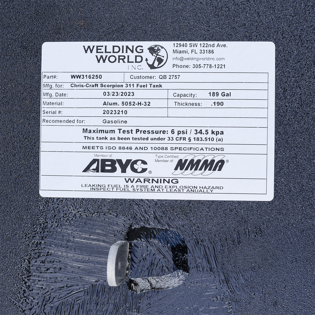 Close-up view of a white rectangular label on a textured black surface. The label is from Welding World Inc., located at 12940 SW 122nd Ave, Miami, FL 33186, with contact info and phone number 305-778-1221. It shows part number WW316250, customer QB 2757, manufacturing for Chris-Craft Scorpion 311 Fuel Tank, manufacturing date 03/23/2023, capacity 189 gallons, material Aluminum 5052-H-32, thickness 0.190, serial number 2023210, recommended for gasoline