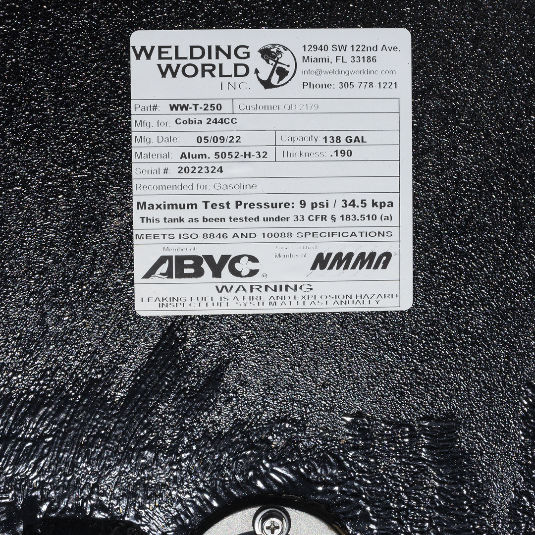A close-up of a white product label from "Welding World Inc." on a textured black surface. The label provides specifications for a fuel tank, including Part#, Mfg. for, Mfg. Date, Material, Serial #, Capacity, and Thickness. It also states the maximum test pressure, certification details, and a warning about leaking fuel
