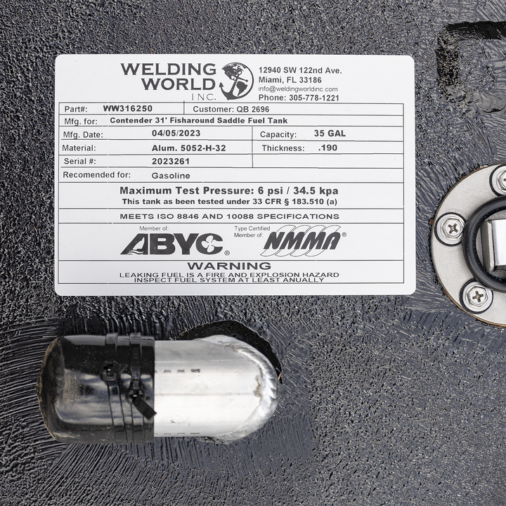Close-up of a white label on a black textured metal surface, showing details from Welding World Inc. The label includes company contact info: 12940 SW 122nd Ave., Miami, FL 33186, info@weldingworldinc.com, Phone: 305-778-1221. Part number WW316250, Customer QB 2696. Manufactured for Contender 31' Fisharound Saddle Fuel Tank. Manufacturing date: 04/05/2023. Capacity: 35 GAL. Material: Aluminum 5052-H-32. Thickness: .190. Serial number: 2023261. Recommended for gasoline