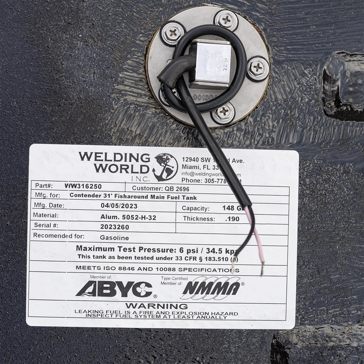 Close-up of a white label attached to a black textured metal surface. The label shows Welding World Inc. company info with address 12940 SW 122nd Ave., Miami, FL 33186 and phone number 305-778-1221. It lists part number VW316250 and customer QB 2696. The label is for a Contender 31' Fisharound Main Fuel Tank, manufactured on 04/05/2023. Capacity is 148 gallons, made of Aluminum 5052-H-32 with thickness 0.190. Serial number 2023260. Recommended for gasoline. Maximum test pressure is 6 psi / 34.5 kpa