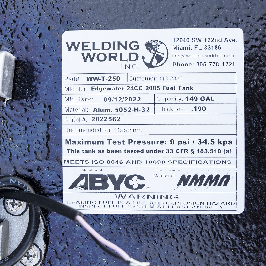 A close-up of a white manufacturer's label on a heavily textured black surface, positioned next to a silver fuel sending unit on the left. The "Welding World, Inc." label identifies the product as an "Edgewater 24CC 2005 Fuel Tank" with a 149-gallon capacity, made on September 12, 2022. A black wire from the sending unit curves across the bottom of the frame. The label also features ABYC and NMMA compliance logos