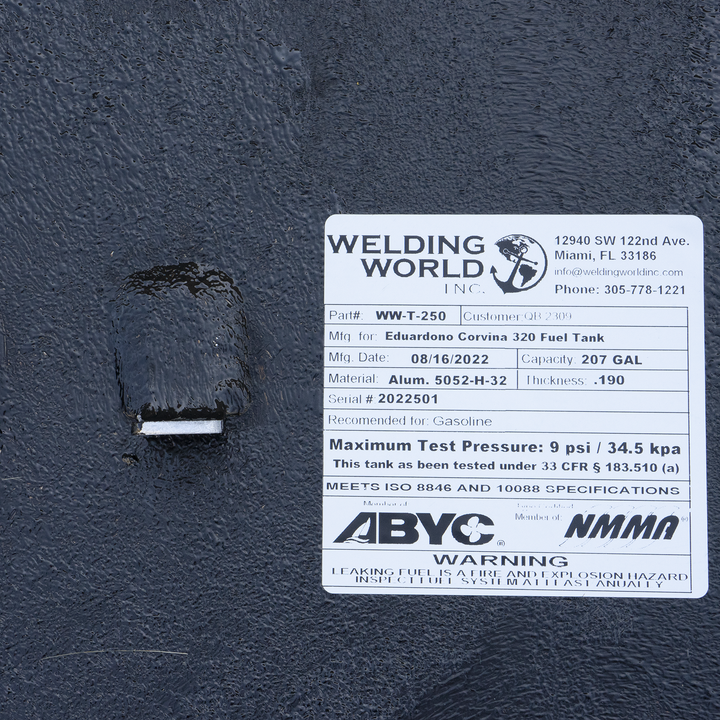 A label on a black fuel tank identifies it as a Welding World Inc. WW-T-250 model, manufactured on August 16, 2022, for an Eduardono Corvina 320 Fuel Tank, with a 207-gallon capacity. It is made of Alum. 5052-H-32 with a thickness of .190, serial number 2022501, and is recommended for gasoline. The label also states a maximum test pressure of 9 psi / 34.5 kpa and that it meets ISO 8846 and 10088 specifications