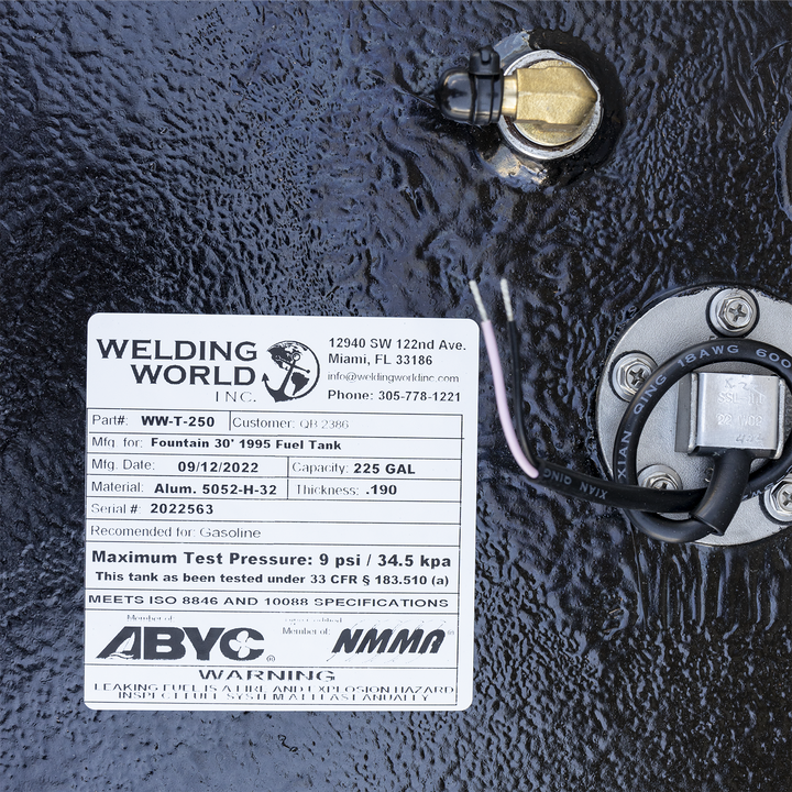 A close-up of a white manufacturer's label on a heavily textured black surface, surrounded by fittings. To the right of the label is a round, silver fuel sending unit, and a brass right-angle fitting is visible above. The "Welding World, Inc." label specifies the tank is for a "Fountain 30' 1995 Fuel Tank" with a 225-gallon capacity, made on September 12, 2022, and features ABYC and NMMA compliance logos