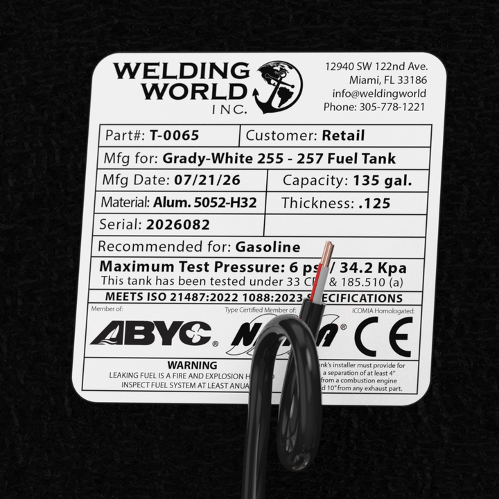 A macro top-down studio view of the top deck hardware cluster and certification label on a physical, finished black encapsulated Grady-White Freedom 255-257 OEM replacement fuel tank (135 gal) from file image_990078.jpg. Positioned against the textured black TarGard epoxy coating, the hardware assembly features a central circular silver KUS electronic fuel level sending unit secured with five bolts and a looped black wire harness. Surrounding fittings include dual pre-installed high-flow brass 90-degree fue
