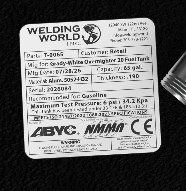A macro top-down view of the official manufacturer certification tag on a physical, finished black encapsulated Grady-White Overnighter 20 OEM replacement fuel tank (65 gal) from file image_5512dd.jpg. Adhered to the textured black TarGard epoxy coating, the weather-resistant specification label from Welding World Inc. (Miami, FL) lists part number T-0065, heavy-gauge Alum. 5052-H32 material, .190 thickness, a 65 gal. capacity, gasoline recommendation, 6 psi / 34.2 Kpa maximum test pressure under 33 CFR § 1
