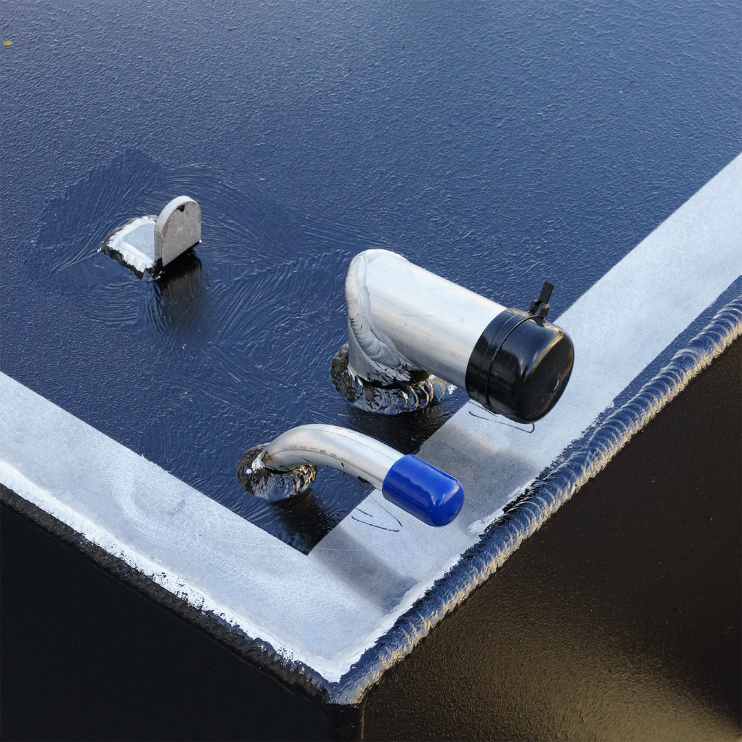 A close-up of a dark, textured surface, likely part of a fuel tank, with a prominent light gray border. Several metal fittings are visible, including a small L-shaped bracket, an angled silver pipe with a black cap, and a curved silver pipe with a blue cap. The surface shows some streaking, possibly from liquid or residue