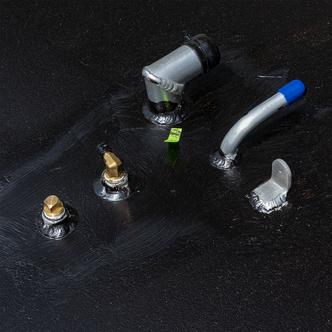 Alt text: Image shows various metal fittings, including angled pipes and brass connectors, on the dark, textured surface of a Mako 23' 1986 - 177 gal. OEM replacement belly fuel tank. A small piece of green tape is visible on the surface