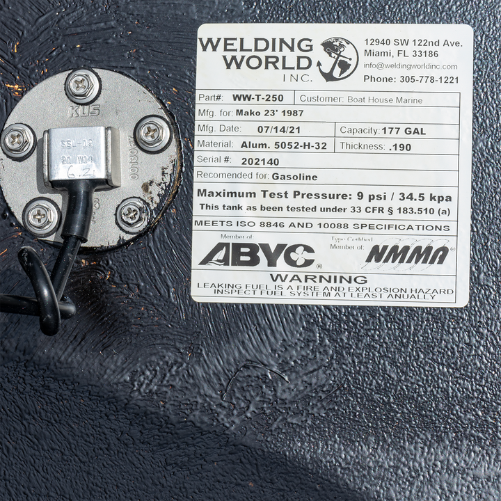 Alt text: A close-up of a fuel tank with a white product label from "Welding World Inc." The label details specifications for a Mako 23' 1987, 177-gallon fuel tank, including material (Alum. 5052-H-32), thickness (.190), serial number (202140), and recommended use for gasoline. It also states the maximum test pressure (9 psi / 34.5 kPa) and compliance with ISO 8846 and 10088 specifications, and ABYC and NMMA certifications. A warning about leaking fuel is also visible