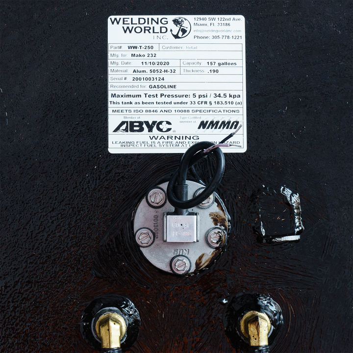 A close-up of a specification label from Welding World Inc., detailing the Mako 232 tank with part number WW-T-250. The label includes manufacturing date, capacity (157 gallons), material specifications, maximum test pressure, and safety warnings regarding leaking fuel hazards. It prominently features compliance certifications and a circular connection with wiring