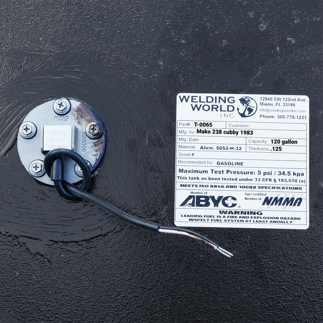 Close-up photograph of a manufacturing certification label for a 120-gallon Mako 238 Cubby (1983) fuel tank by Welding World Inc., specifying .125" thickness and 5052-H32 aluminum, shown next to a KUS fuel sending unit on a textured coal-tar epoxy finish.