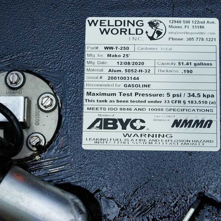 A close-up shot of a white rectangular product label from "Welding World Inc." affixed to a textured black surface. The label provides details for a fuel tank: Part# WW-T-250, manufactured for Mako 25' on 12/08/2020, with a capacity of 51.41 gallons. The material is Alum. 5052-H-32 with a thickness of .190, serial # 2001003144, and it's recommended for gasoline. It specifies a maximum test pressure of 5 psi / 34.5 kpa and notes compliance with ISO 8846 and 10088 specifications, with membership in ABYC/NMMA
