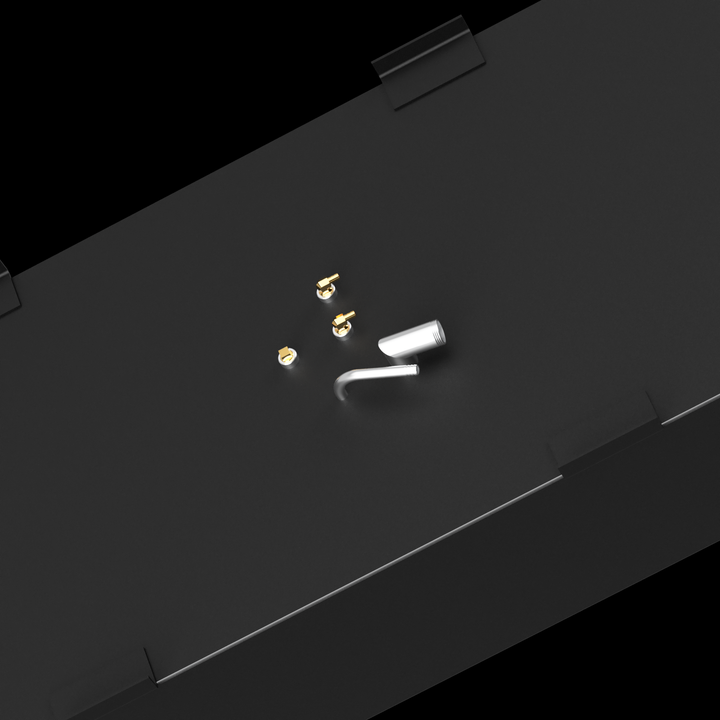 A close-up, angled view of the top surface of a dark grey fuel tank against a black background. Several golden-colored threaded fittings and two white, curved pipe sections are visible on the surface. Black L-shaped brackets are present along the top edges of the tank