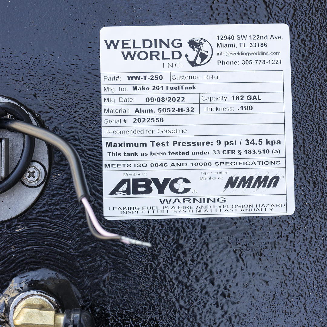 A close-up of a white manufacturer's label on a heavily textured black surface, situated between a fuel sending unit on the left and a brass fitting below. The "Welding World, Inc." label identifies the product as a "Mako 261 Fuel Tank" with a 182-gallon capacity, made on September 8, 2022. A black wire from the sending unit curves across the bottom of the frame. The label also features ABYC and NMMA compliance logos