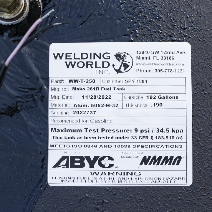 A close-up photograph of a white manufacturer's label on a black fuel tank. The label is for "Welding World, Inc." and details the specifications for a "Mako 261B Fuel Tank." Key information listed includes a 192-gallon capacity, an aluminum material type, a manufacturing date of 11/28/2022, and a serial number. The label also displays compliance logos for ABYC and NMMA, pressure testing data, and a safety warning at the bottom. The edge of a fuel fitting and a wire are visible in the upper left corner.