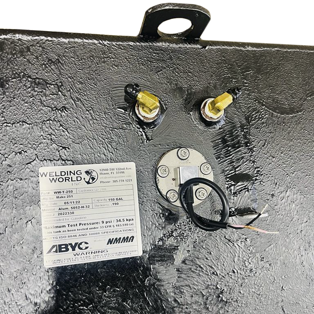 An overhead close-up view of a textured dark grey surface, likely a fuel tank, shows a white product label from "WELDING WORLD INC." detailing product specifications for a "Mako 251" fuel tank with a 150-gallon capacity. Above the label, two brass fittings with black caps are visible. A circular metallic plate secured by screws with a black wire extending from its center is also present. In the upper left corner, a black metal handle or loop is partially visible