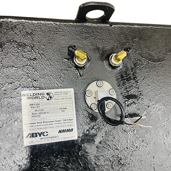An overhead close-up view of a textured dark grey surface, likely a fuel tank, shows a white product label from "WELDING WORLD INC." detailing product specifications for a "Mako 251" fuel tank with a 150-gallon capacity. Above the label, two brass fittings with black caps are visible. A circular metallic plate secured by screws with a black wire extending from its center is also present. In the upper left corner, a black metal handle or loop is partially visible