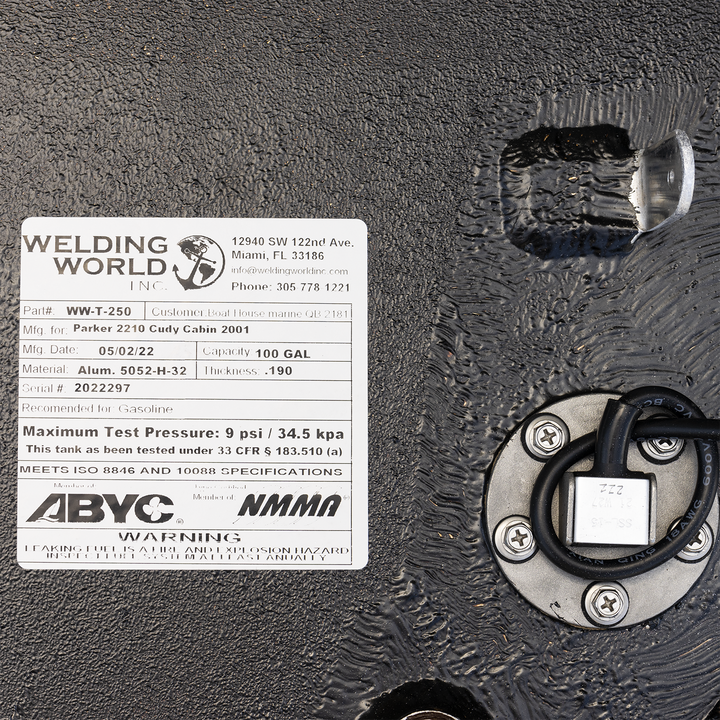 A close-up of a white product label for a "WW-T-250" fuel tank manufactured by Welding World Inc. for a "Parker 2210 Cuddy Cabin 2001". The label details the tank's capacity (100 gal), material (Alum. 5052-H-32), thickness (.190), and manufacturing date (05/02/22). It also specifies a maximum test pressure and states compliance with ISO and NMMA standards. The label is attached to a textured black surface, and part of a circular sensor with wires is visible on the right