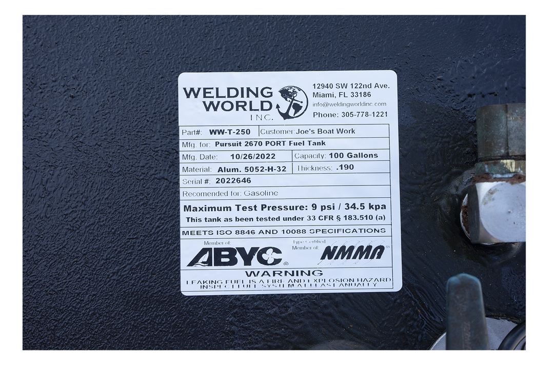 A close-up photograph of a white manufacturer's label on a black, textured surface. The label from "Welding World, Inc." identifies the item as a "Pursuit 2670 STBD Fuel Tank" with a 100-gallon capacity, manufactured on October 26, 2022. Compliance logos for ABYC and NMMA are displayed above a safety warning at the bottom of the label.

