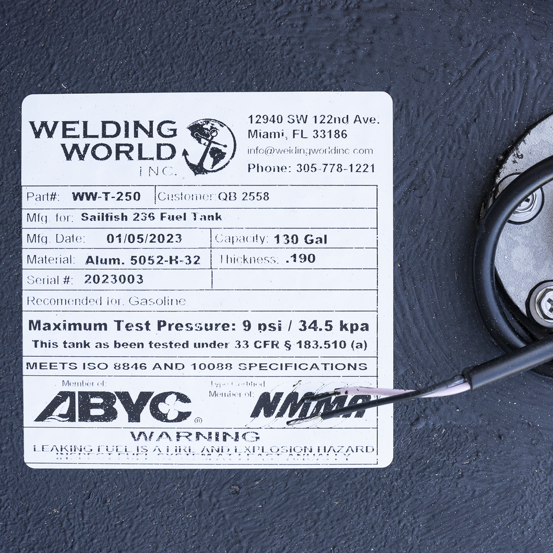 A close-up photo of a white label on a textured black surface showing product information from Welding World Inc. The label indicates the part number WW-T-250 for a Sailfish 236 Fuel Tank manufactured on 01/05/2023. The tank has a capacity of 130 gallons and is made of aluminum 5052-K-32 with a thickness of .190 inches. The serial number is 2023003, and the tank is recommended for gasoline use. The label also notes a maximum test pressure of 9 psi / 34.5 kpa and compliance with ISO 8846 and 10088