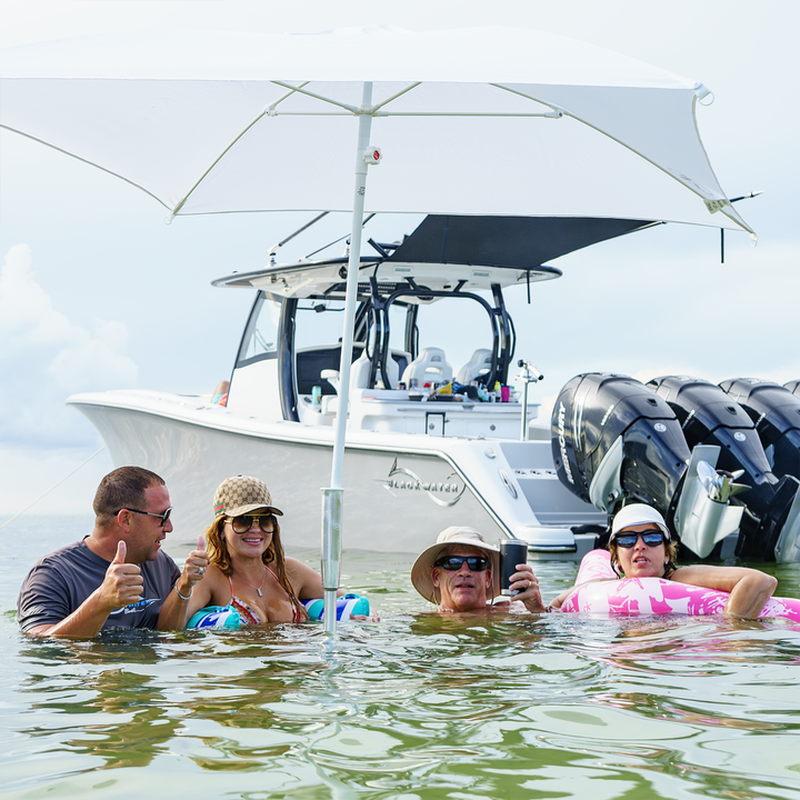 A group of people enjoying a day at the sandbar, giving thumbs up while standing in shallow water. In the background, a boat with multiple outboard motors is anchored under a large umbrella, providing shade. The scene captures a relaxed and fun atmosphere on a sunny day