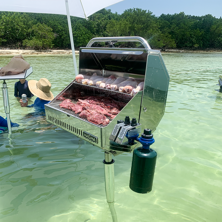 A stainless steel grill mounted on a pole, standing in shallow water with various meats cooking on the grill. In the background, lush green trees line the shore, and people in sun hats are enjoying the beach atmosphere. A propane tank is attached to the grill, and some individuals are holding drinks while wading in the water