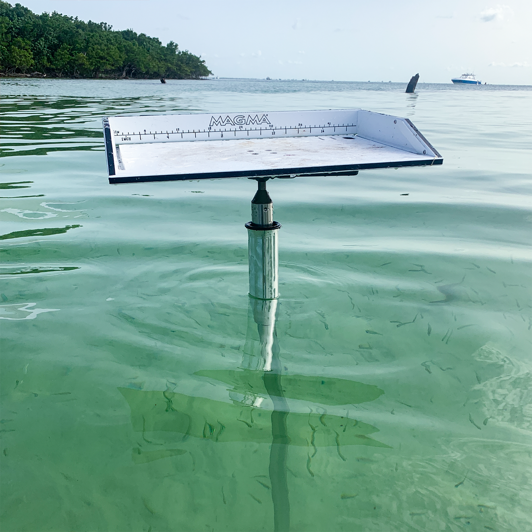 A sandbar spike is partially submerged in clear, shallow water, with a flat, rectangular measuring surface marked with measurements. The spike is mounted on a vertical pole, and the surrounding water is calm, reflecting the sky and distant greenery. In the background, a boat can be seen on the horizon.