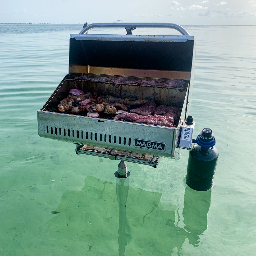 A Magma grill mounted on a pole stands in shallow, clear water, with various meats and vegetables sizzling on the grill. The scene captures a serene outdoor cooking experience, with a calm sea and a clear sky in the background