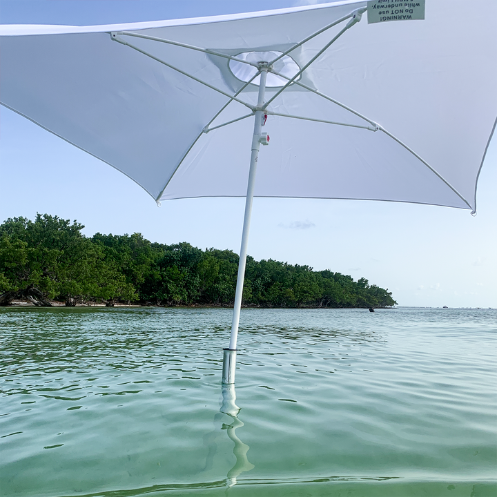 A white beach umbrella stands upright in shallow, clear water, with its pole anchored in the sand below. In the background, lush green trees line the shore under a clear blue sky. The water reflects the umbrella and the surrounding scenery, creating a serene beach atmosphere