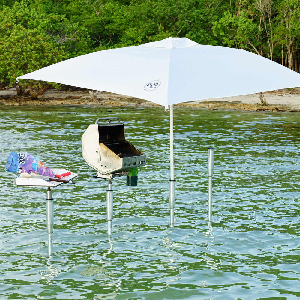 A grill set up on a sandbar in shallow water, supported by metal poles. A large white umbrella provides shade over the grill, while a small table nearby holds food items, including a pack of hot dogs and a plate. The background features lush greenery along the shoreline