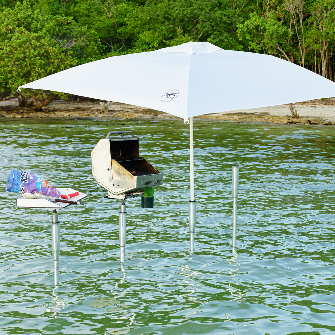 A grill set up on a sandbar in shallow water, supported by metal poles. A large white umbrella provides shade over the grill, while a small table nearby holds food items, including a pack of hot dogs and a plate. The background features lush greenery along the shoreline