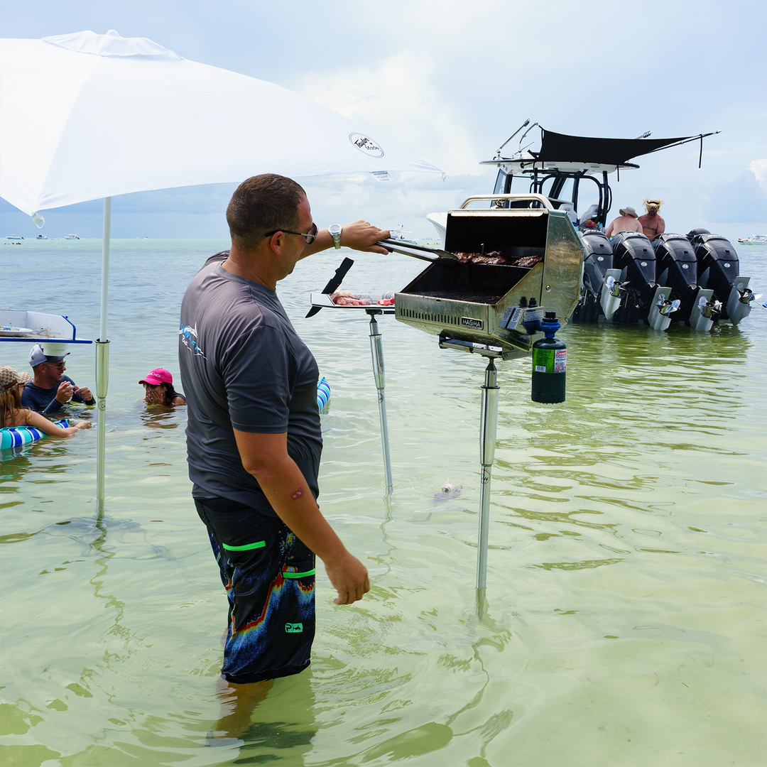 A man stands in shallow water, grilling food on a portable grill attached to a boat. He is wearing a gray shirt and colorful swim trunks, with a large umbrella providing shade. In the background, several people are enjoying the water, and a boat with multiple outboard motors is anchored nearby. The scene captures a relaxed day at a sandbar