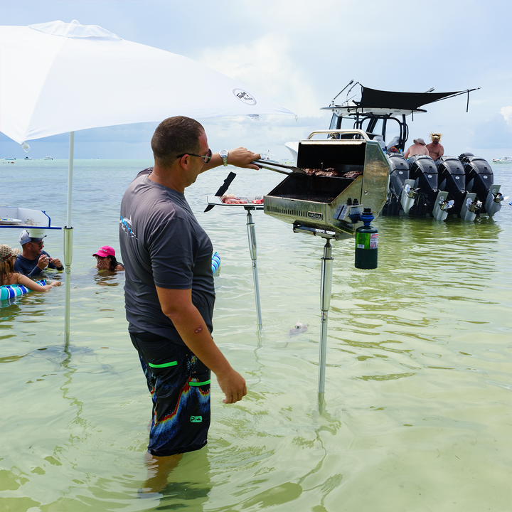 A man stands in shallow water, grilling food on a portable grill attached to a boat. He is wearing a gray shirt and colorful swim trunks, with a large umbrella providing shade. In the background, several people are enjoying the water, and a boat with multiple outboard motors is anchored nearby. The scene captures a relaxed day at a sandbar
