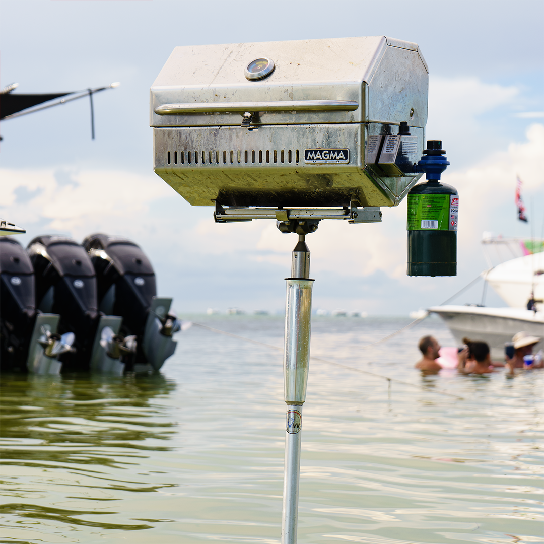 Alt text: A stainless steel Magma grill mounted on a pole near the water, with several boats in the background and people enjoying a day at the beach. The sky is partly cloudy, and the grill is ready for use