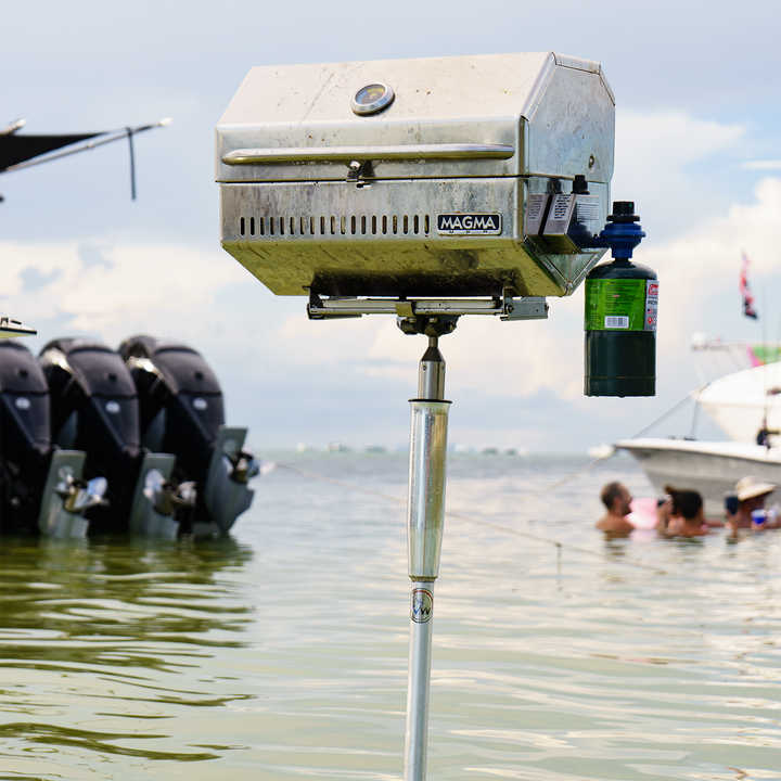 Alt text: A stainless steel Magma grill mounted on a pole near the water, with several boats in the background and people enjoying a day at the beach. The sky is partly cloudy, and the grill is ready for use