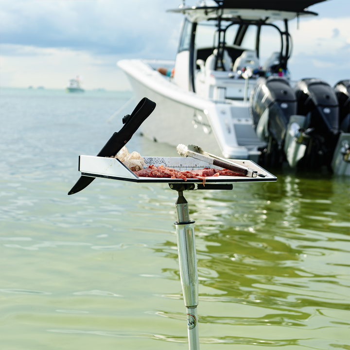 A portable grill set up on a sandbar, with various meats cooking on it. In the background, a boat with multiple outboard motors is anchored in shallow water, surrounded by a calm sea