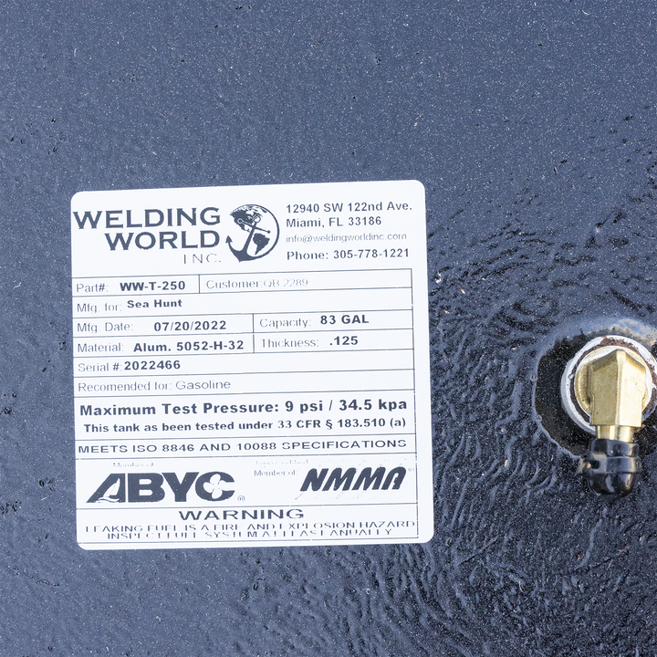 A long, rectangular black fuel tank with a slightly rounded front end, viewed from above and at an angle. The top surface is wet and reflective, showing various metal fittings and connections, including a white and blue pipe assembly on the right and a circular fitting with multiple small pipes on the left. Four dark metal brackets are attached to the top edges of the tank