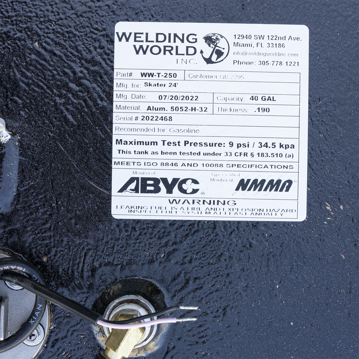 A white product label from "WELDING WORLD INC." is affixed to a textured black surface, likely a fuel tank. The label details the tank's specifications: Part# WW-T-250, manufactured for "Skater 24'," Mfg. Date 07/20/2022, Capacity 40 GAL, Material Alum. 5052-H-32, Thickness .190, and Serial # 2022468. It is recommended for gasoline and has a maximum test pressure of 9 psi / 34.5 kpa. The tank meets ISO 8846 and 10088 specifications and is certified by ABYC and NMMA
