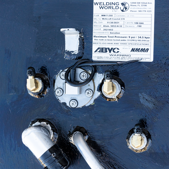 A close-up view of a black, textured surface, likely the top of a fuel tank, with various metal fittings, connectors, and a white label from "WELDING WORLD INC." providing specifications like part number, manufacturing details, capacity (188 GAL), material (Alum. 5052-H-32), and a warning about leaking fuel